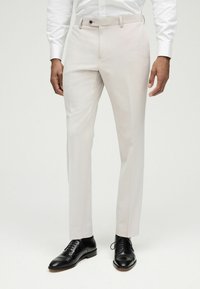 Pantalones de vestir color beige claro con un corte ajustado, parte frontal plana, pasadores para cinturón y cierre de un solo botón. Combinados con zapatos de vestir de piel negra.
