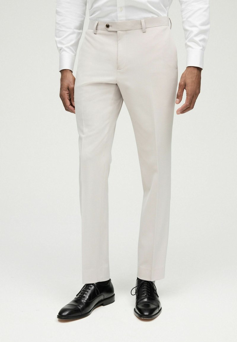Pantalones de vestir color beige claro con un corte ajustado, parte frontal plana, pasadores para cinturón y cierre de un solo botón. Combinados con zapatos de vestir de piel negra.
