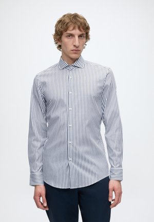 Homme portant une chemise blanche à manches longues à rayures verticales noires et un pantalon sombre, debout sur un fond blanc.