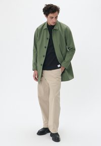Chaqueta ligera verde con cierre de botones y cuello puntiagudo, llevada sobre una camisa negra y pantalones beiges; presenta un diseño minimalista.