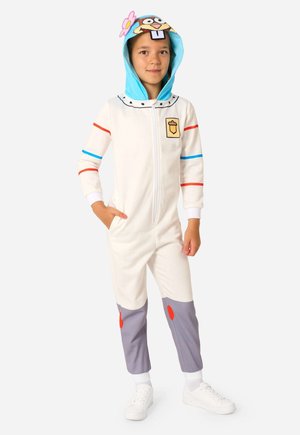 Kind draagt een wit jumpsuit met capuchon met kleurrijke strepen en een karaktergezicht op de capuchon, staat met één hand in de zak en witte sneakers.