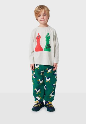 Langärmliges graues Shirt mit roten und grünen Schachfiguren-Grafiken, kombiniert mit grünen Hosen mit Entenmuster und gelben Schnürsenkeln an den Schuhen.