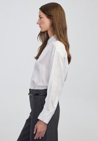 Weiße Bluse mit langen Ärmeln und dünnen vertikalen Streifen, mit Stehkragen und weiten Ärmeln, kombiniert mit dunklen, maßgeschneiderten Hosen.