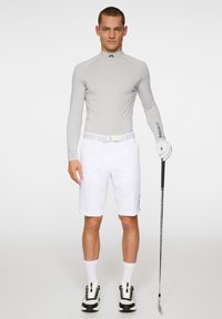 Maglietta grigia chiara a maniche lunghe, pantaloni corti bianchi con cintura, calze bianche, scarpe nere e bianche, tiene in mano un bastone da golf. Design semplice e aderente.