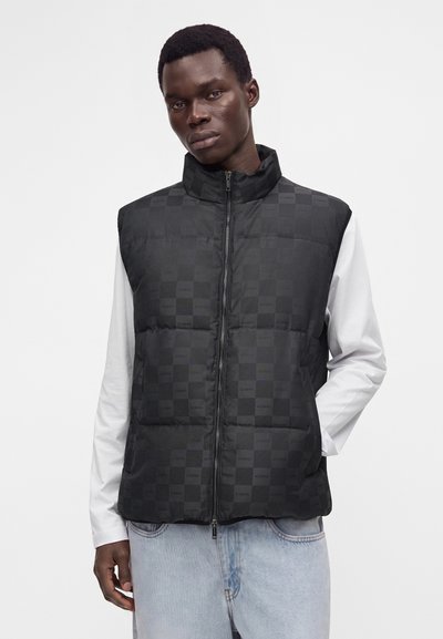 Homme portant un gilet matelassé noir avec un motif à carreaux subtil sur une chemise blanche à manches longues et un jean bleu clair, devant un fond uni.