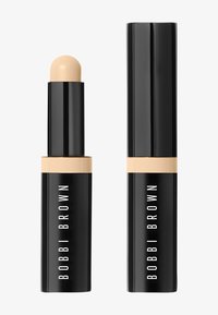 Correttore in stick Bobbi Brown, con involucro nero e dettagli dorati. Parte superiore rotonda, texture liscia, colore crema beige, etichettato con il nome del marchio.