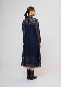 Tmavomodré čipkované šaty s dlhými rukávmi a vysokým golierom, so priehľadným dizajnom a kvetinovými vzormi, doplnené čiernymi kovbojskými čižmami.