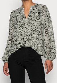 Ljusgrön blus i genomskinlig tyg med V-ringning, långa puffärmar och svarta abstrakta fläckar. Matchas med svarta tajta byxor.