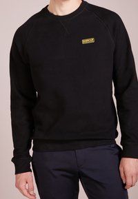 Black sweat shirt featuring a classic crew neck design, ribbed cuffs, and a small yellow "Barbour International" logo on the chest.
