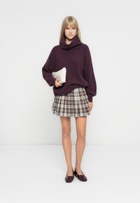Pull en tricot violet à col bénitier, associé à une mini-jupe écossaise marron et crème. Il est également accompagné de ballerines marron et d'une pochette blanche.