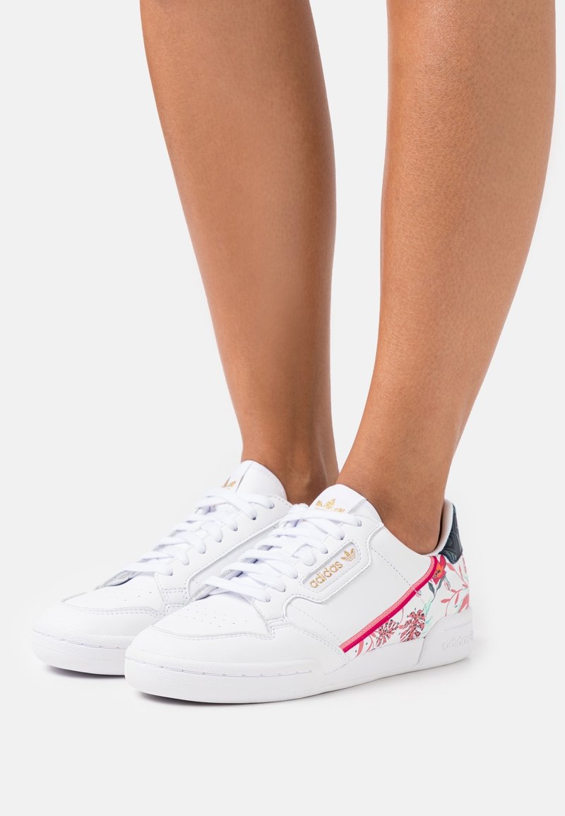 Vita Adidas-sneakers med blommönster och gyllene logotypdetaljer, bärs på bara ben mot en vit bakgrund.