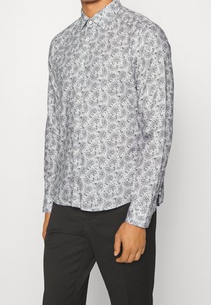 Mężczyzna w białej koszuli na guziki z szarym wzorem paisley i czarnych spodniach, stojący na białym tle.