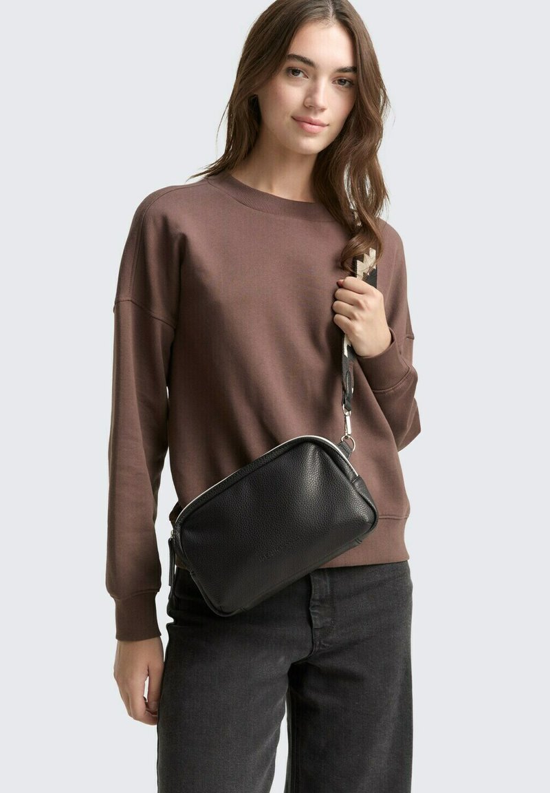 Borsa a tracolla in pelle nera con superficie texture, hardware in argento e una tracolla a fantasia, indossata con una felpa marrone e jeans grigi.