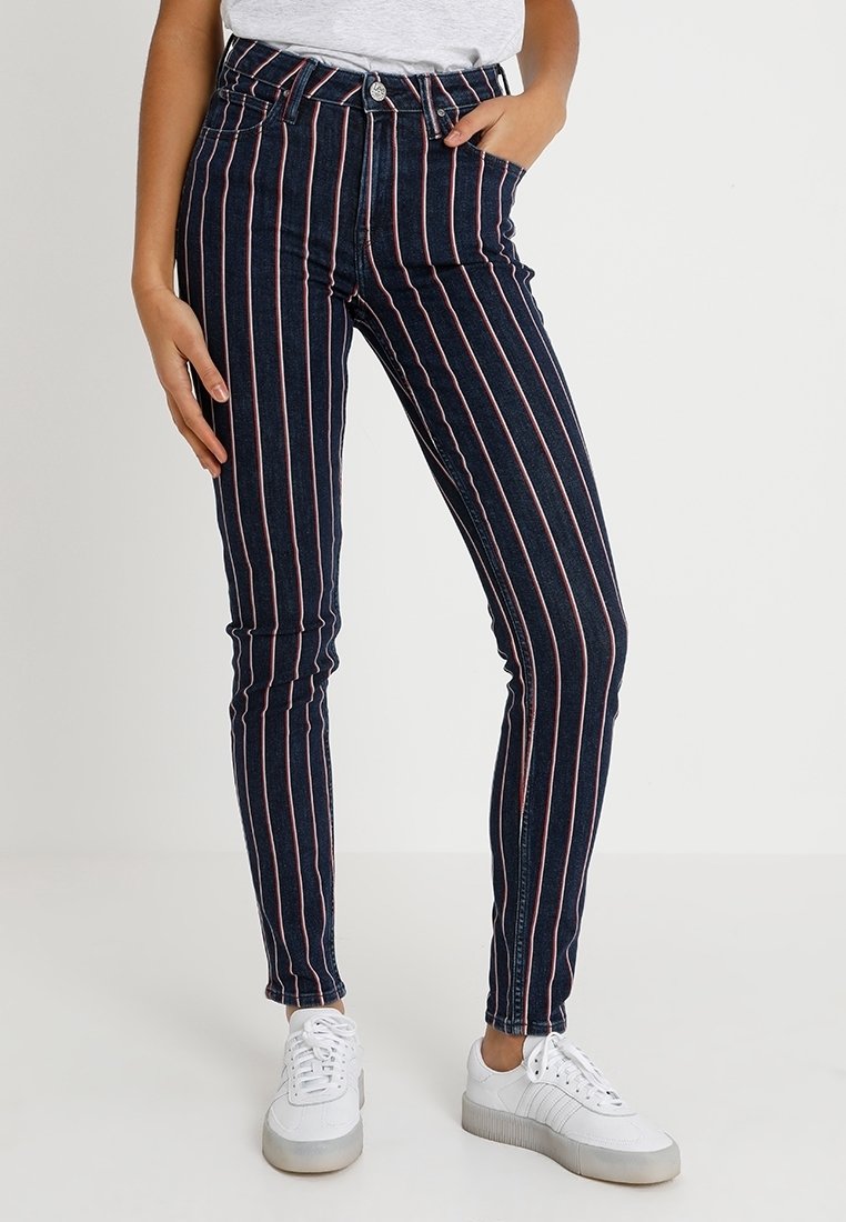 Jeans de tiro alto en color azul marino con rayas verticales rojas, con un diseño ajustado y estilo clásico de cinco bolsillos, combinados con zapatillas blancas.