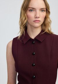 Vestito senza maniche color borgogna con scollo a colletto, caratterizzato da sette bottoni neri sul davanti e una texture del tessuto liscia.