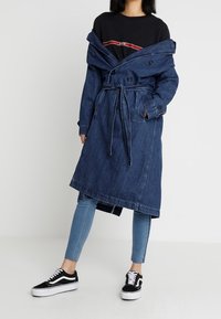 Trench-coat en jean avec une ceinture à la taille, de gros boutons et des revers, porté par-dessus un t-shirt noir et associé à un jean bleu clair et des baskets noires.