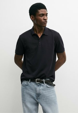 Jonge man draagt een zwart poloshirt met korte mouwen dat in een lichtblauwe spijkerbroek is gestopt met een zwarte gevlochten riem, staand met zijn handen achter zijn rug.