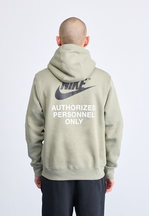 Personne au crâne rasé portant un sweat à capuche Nike vert olive clair avec l'inscription « PERSONNEL AUTORISÉ SEULEMENT » au dos, debout devant un fond uni.