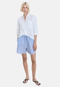 Chemise en lin blanche, shorts rayés bleus avec taille élastique et cordon de serrage, et tongs noires avec des pois blancs.