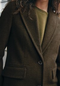 Blazer en laine vert olive avec revers crantés, deux poches avant et boutons foncés. Le tissu texturé offre une apparence structurée.