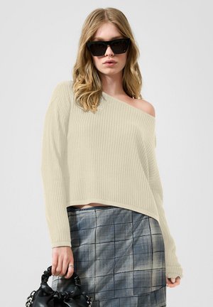 Jeune femme portant un pull beige épaules dénudées, une jupe à carreaux, de grandes lunettes de soleil noires et tenant un sac à main noir sur un fond uni.