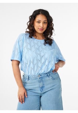 Giovane donna con capelli ricci che indossa un top a maniche corte azzurro chiaro con fantasia e jeans azzurri a vita alta, posando con la mano sul fianco.