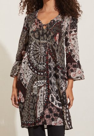 Femme portant une robe à manches longues avec des motifs patchwork en noir, blanc, rouge et motifs floraux, avec un nœud au niveau du col.