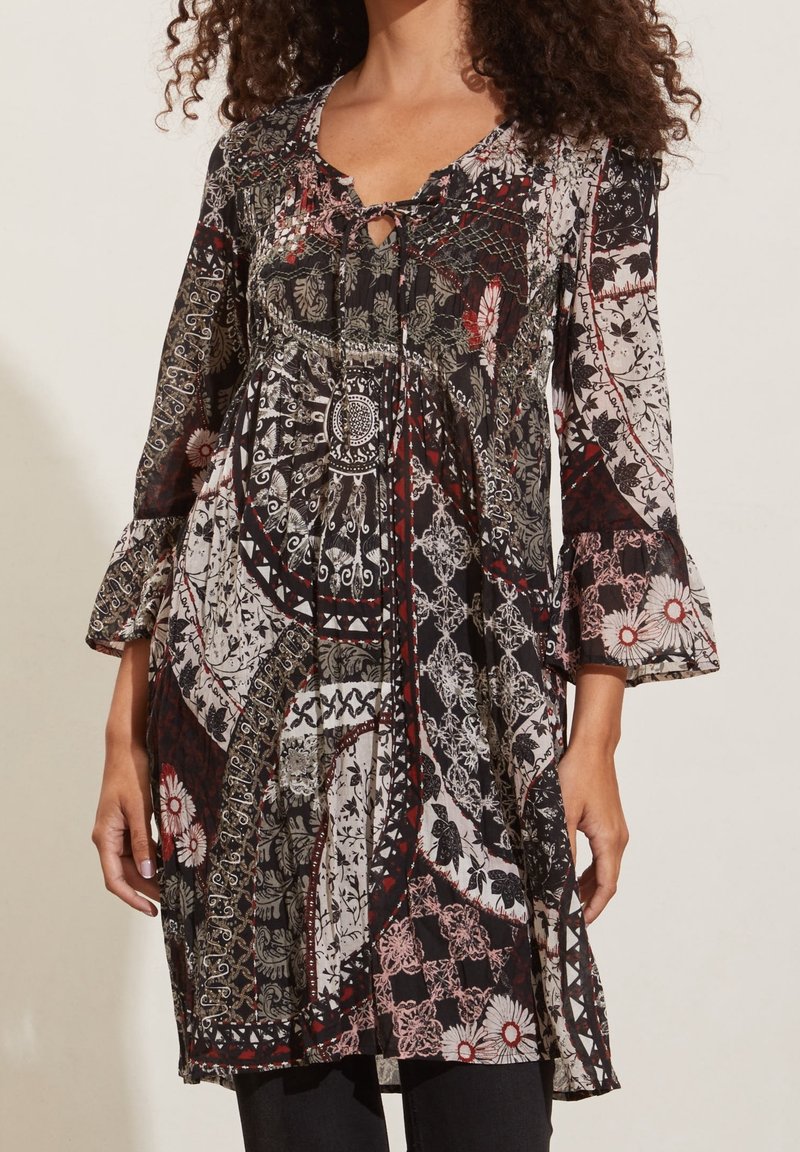 Femme portant une robe à manches longues avec des motifs patchwork en noir, blanc, rouge et motifs floraux, avec un nœud au niveau du col.