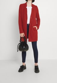 Rode mantel met lange mouwen en knoopsluiting, gedragen over een wit shirt en donkere jeans. Zwarte emmer tas en zwarte schoenen met dubbele banden.