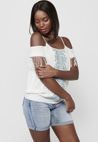 Top bianco scollato con ricami turchesi e maniche frangiate, abbinato a pantaloni corti in denim chiaro. Tessuto morbido e vestibilità rilassata.