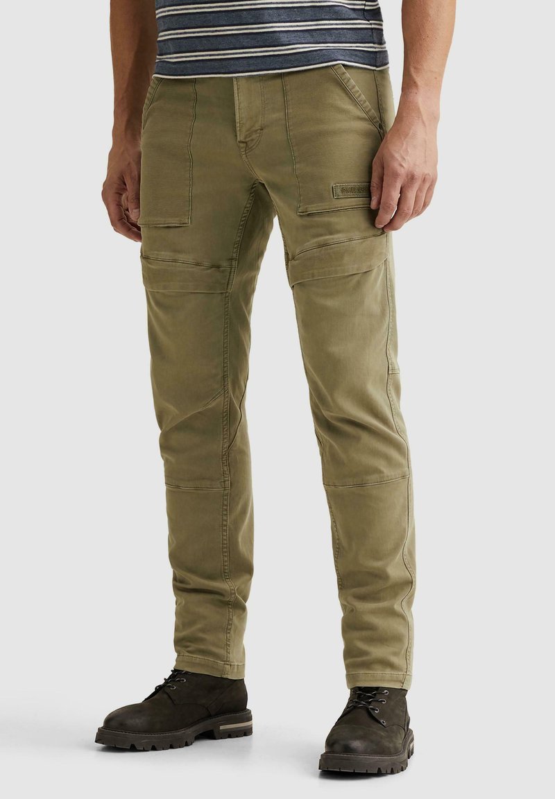 Homme portant un pantalon cargo vert olive et des bottes noires à lacets, debout devant un fond clair uni.