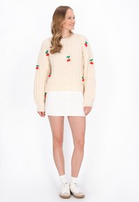 Pletený krémový sveter s červenými a zelenými výšivkami čerešní, skombinovaný s bielou mini sukňou a bielymi teniskami s béžovými akcentmi.