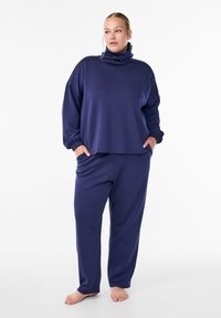 Donna in piedi a piedi nudi che indossa un ampio maglione a collo alto blu navy e pantaloni abbinati, con le mani nelle tasche, su sfondo bianco.