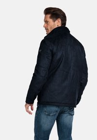 Donkerblauwe suède jas met een hoge kraag, knoopaccenten en een gestructureerd oppervlak, gecombineerd met lichtblauwe denim jeans.