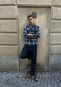 Ung man i mörka jeans och grön rutig skjorta står med armarna i kors och lutar sig mot en beige stenmur på en kullerstensgata.