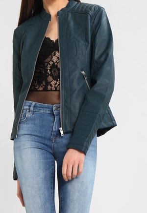Vrouw draagt een donker groene leren jas met rits over een zwart kanten topje, gecombineerd met lichtblauwe high-waisted jeans.