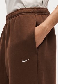 Άτομο που φοράει καφέ φόρμα Nike με ελαστική μέση, χέρι στην πλαϊνή τσέπη και μέρος ανοιχτόχρωμου πουκάμισου ορατό πάνω από τη μέση.