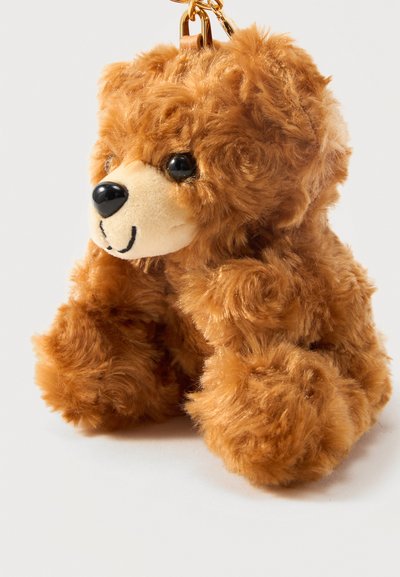 Petit ours en peluche marron avec yeux et nez noirs attaché à un mousqueton doré sur fond blanc.