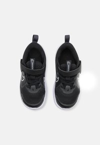 Chaussures de sport noires en tissu en maille, surface texturée, bande velcro et semelle blanche. Embout arrondi et tirette à l'arrière.