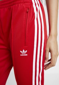 Röda sportbyxor med vita sidostraper och ficka med dragkedja. Har Adidas-logotyp i vitt. Tillverkade av ett mjukt, lätt material.