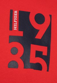 Punainen kangas, jossa on suuri musta painatus numeroista "1985" ja sanasta "HILFIGER" valkoisena, keskitettynä ja modernilla, paksulla fontilla.