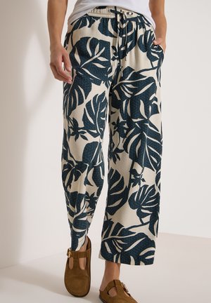 Mujer con pantalones anchos beige con estampado grande de hojas tropicales en azul oscuro y sandalias cerradas marrones con correas y hebillas.