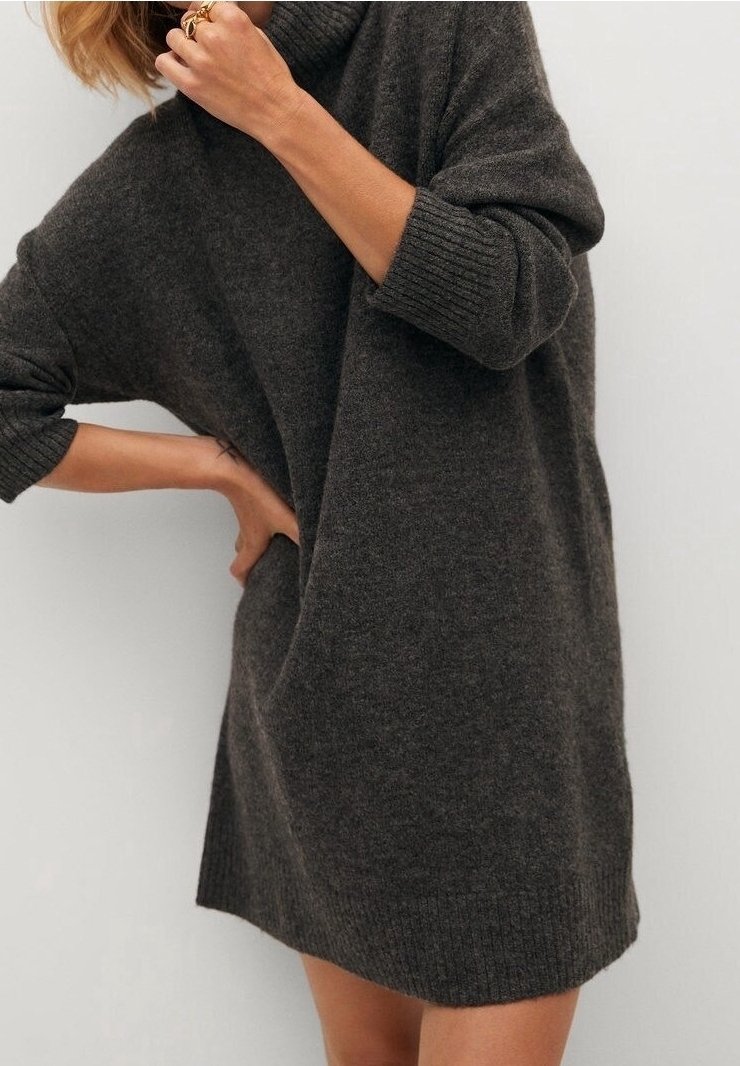 Personne portant une robe-pull à col roulé ample gris foncé avec manches longues, une main sur la hanche et l'autre près du cou montrant des bagues.
