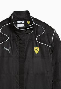 Jachetă neagră din material texturat cu accente albe. Prezintă un guler înalt, un logo argintiu Puma și un emblemă galbenă Ferrari.