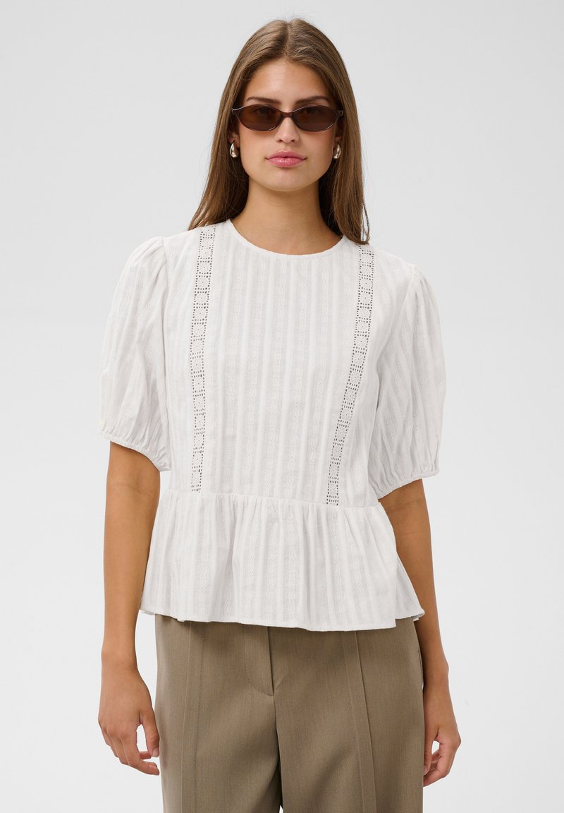 Femme portant des lunettes de soleil ovales marron, des boucles d'oreilles argentées en anneau, un chemisier blanc texturé à manches bouffantes avec des détails en dentelle, et un pantalon beige, fond neutre.