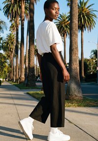 Giovane con camicia bianca, pantaloni neri e sneakers bianche che cammina su un marciapiede fiancheggiato da palme sotto un cielo limpido.