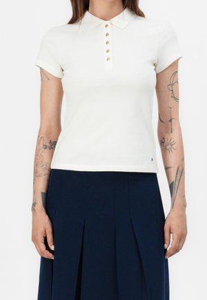 Vrouw die een wit poloshirt met korte mouwen en gouden knopen draagt, en een marineblauwe geplooide rok, zichtbaar van hals tot midden van de dij.