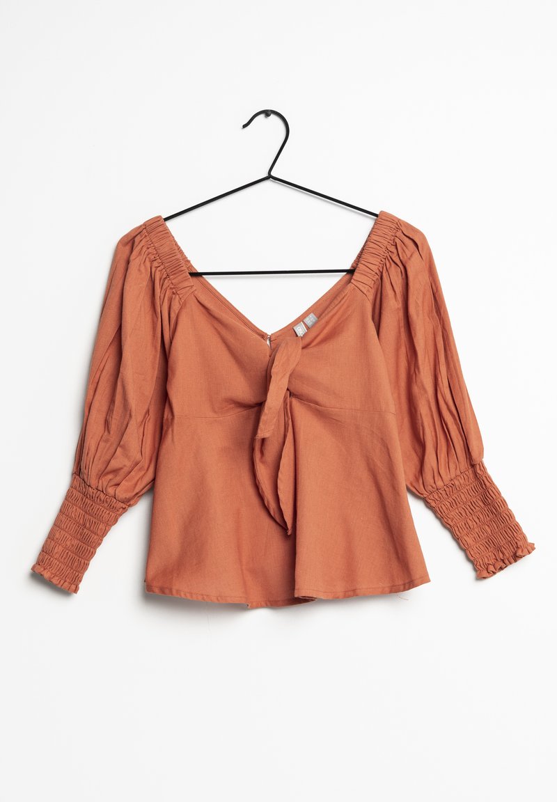 Blouse couleur rouille avec décolleté en V et devant noué. Dotée de manches bouffantes avec des poignets smockés, fabriquée en tissu texturé.
