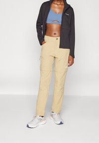 Giacca nera, top corto azzurro chiaro e pantaloni beige con zip. Tessuto liscio e vestibilità rilassata. Sneakers sportive con colori e texture miste.