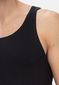 Nahaufnahme einer Person in einem schwarzen ärmellosen Oberteil, das Schulter, Oberarm und teilweise den Brustbereich auf einem schlichten Hintergrund zeigt.